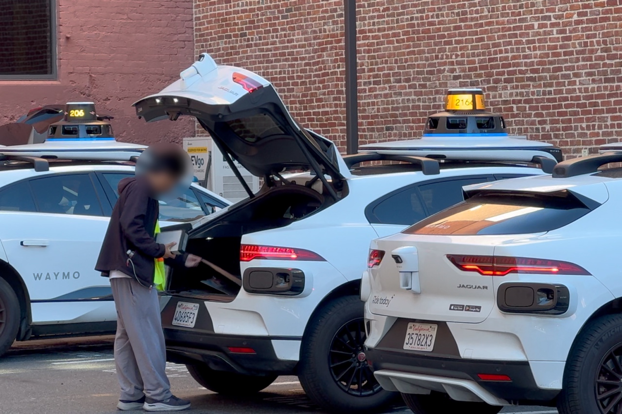 A person installing a gray metallic box in the trunk of a white autonomous vehicle