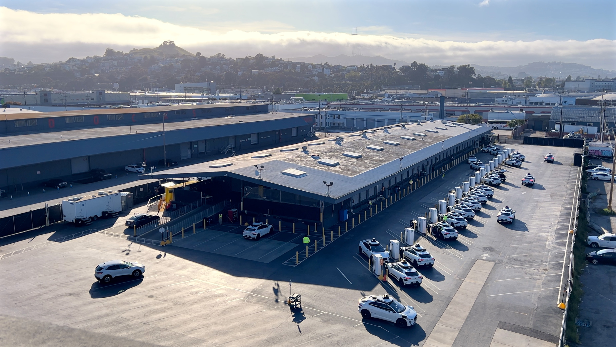 A photograph of a parking lot with white autonomous vehicles and charging equipment in front of a hill on a partly cloudy day