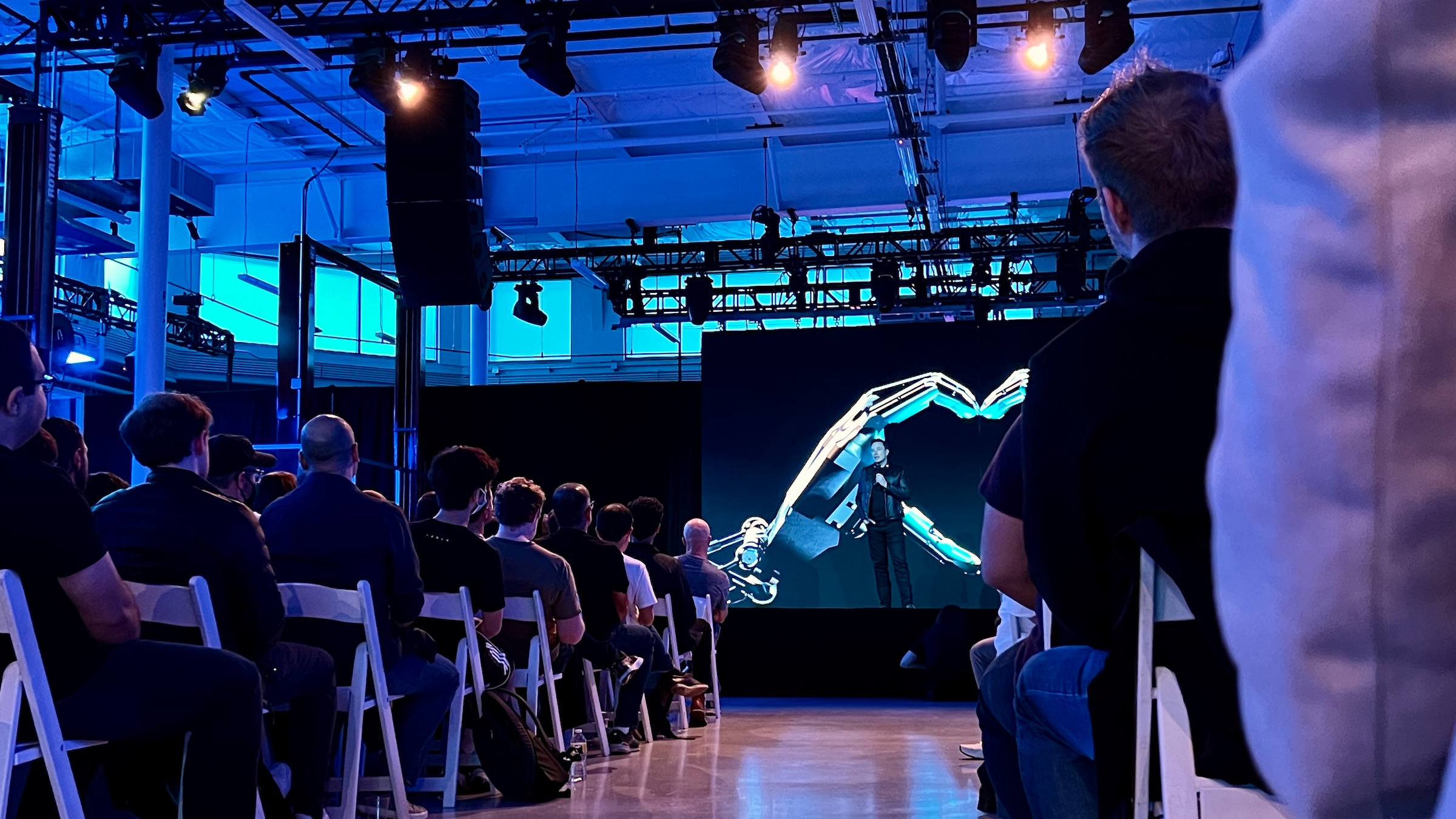 Elon Musk stands on a stage in front of a screen. He addresses a seated audience.